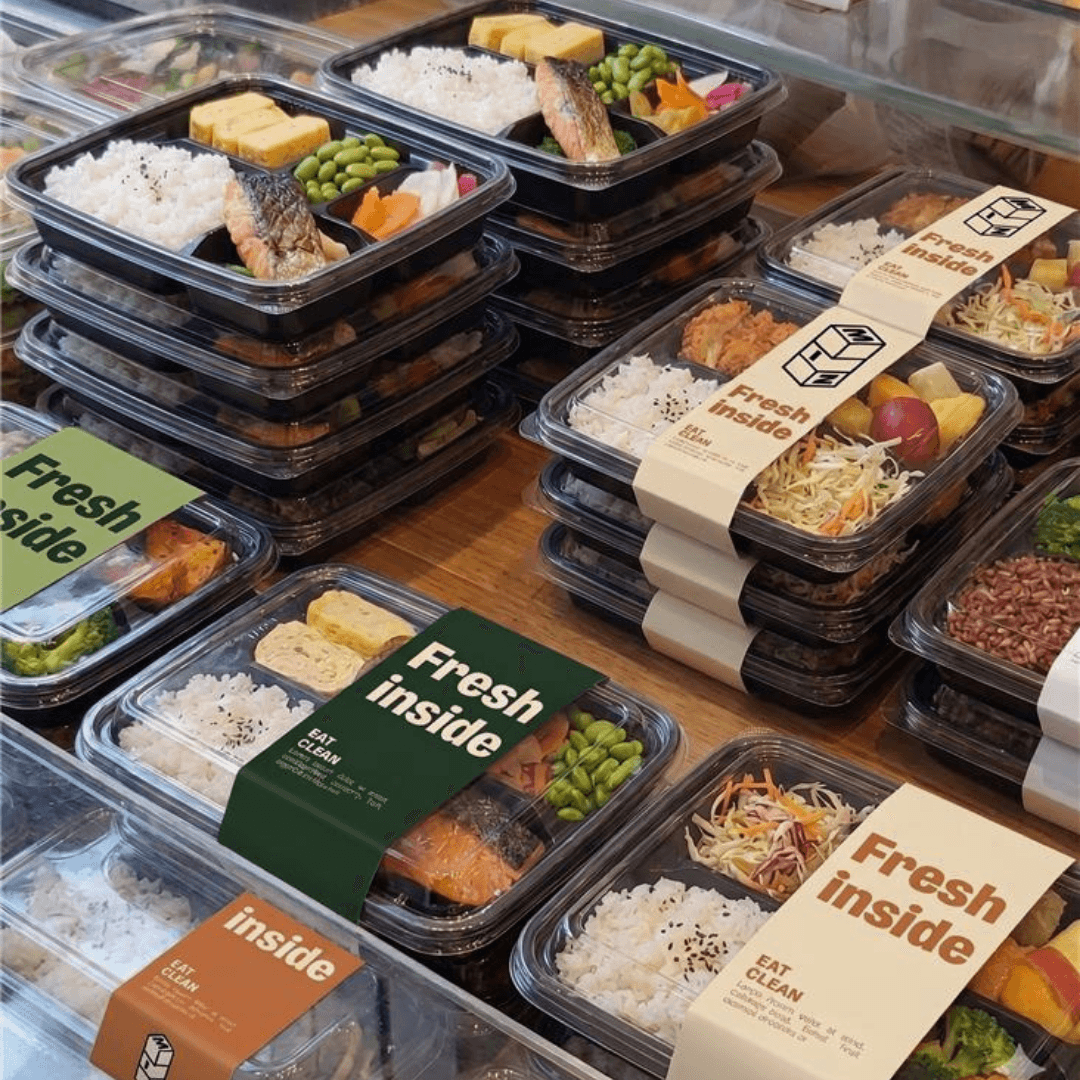 Stacks of fresh prepared meal boxes arranged on a wooden table, featuring balanced grab-and-go meals with rice, vegetables, fresh fruit, and protein-packed entrées in branded packaging.