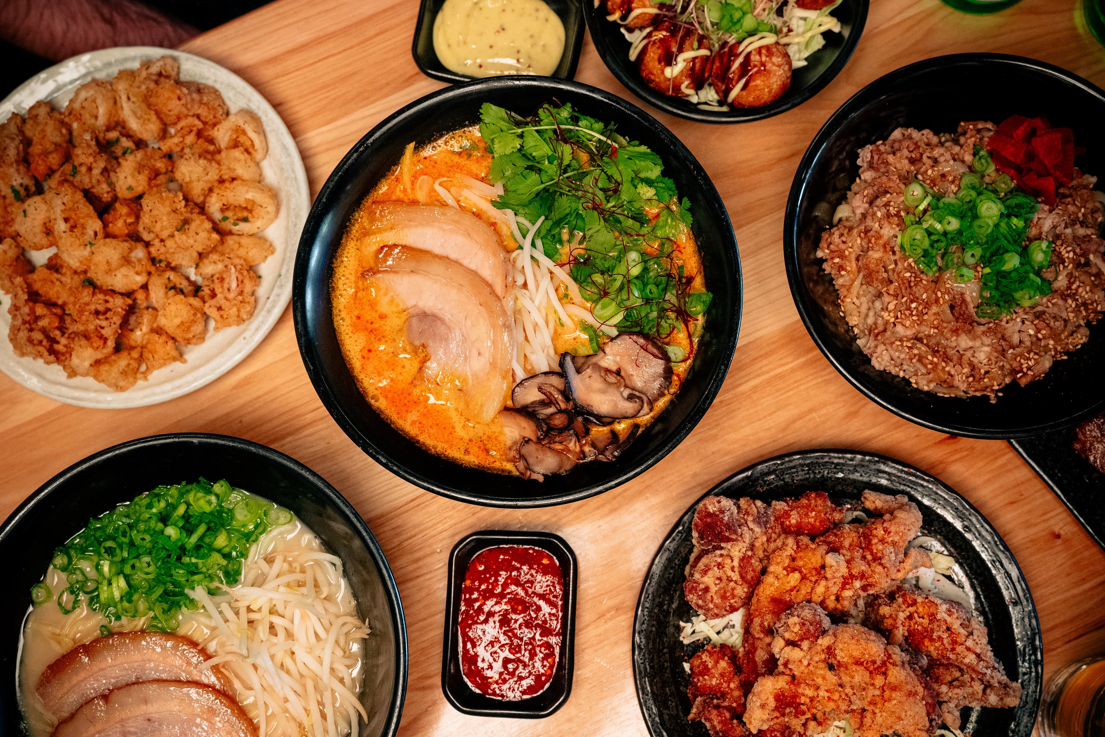 An overhead spread from KIN Craft Ramen & Izakaya featuring chicken karaage, crispy calamari, takoyaki topped with sauce and bonito flakes, fried rice, and a variety of ramen bowls with chashu pork, mushrooms, bean sprouts, and fresh green onions.