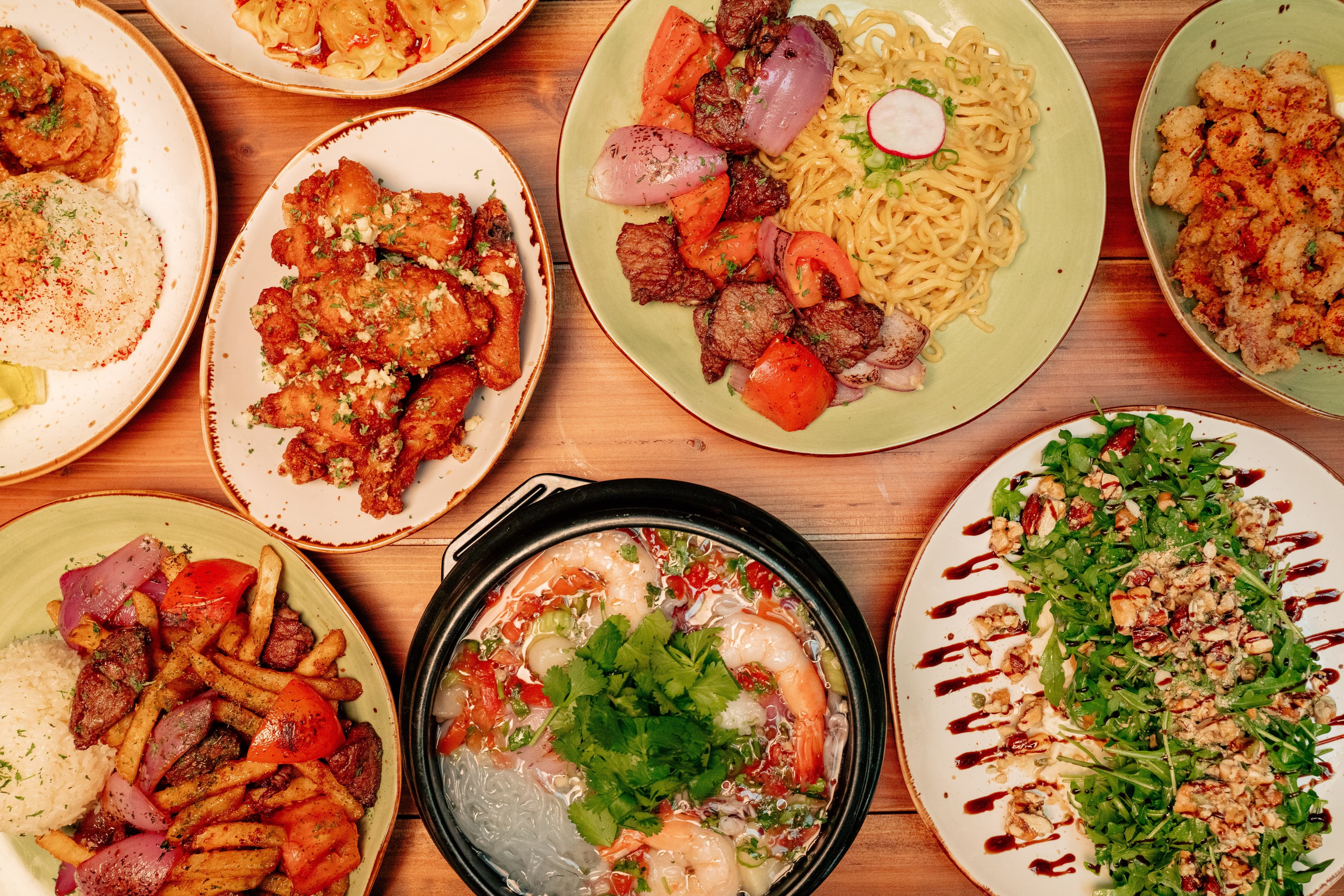 An overhead spread of VOX Kitchen signature dishes featuring filet mignon lomo saltado, seafood soup, crispy chicken wings, calamari, and pear salad arranged across a wooden table.