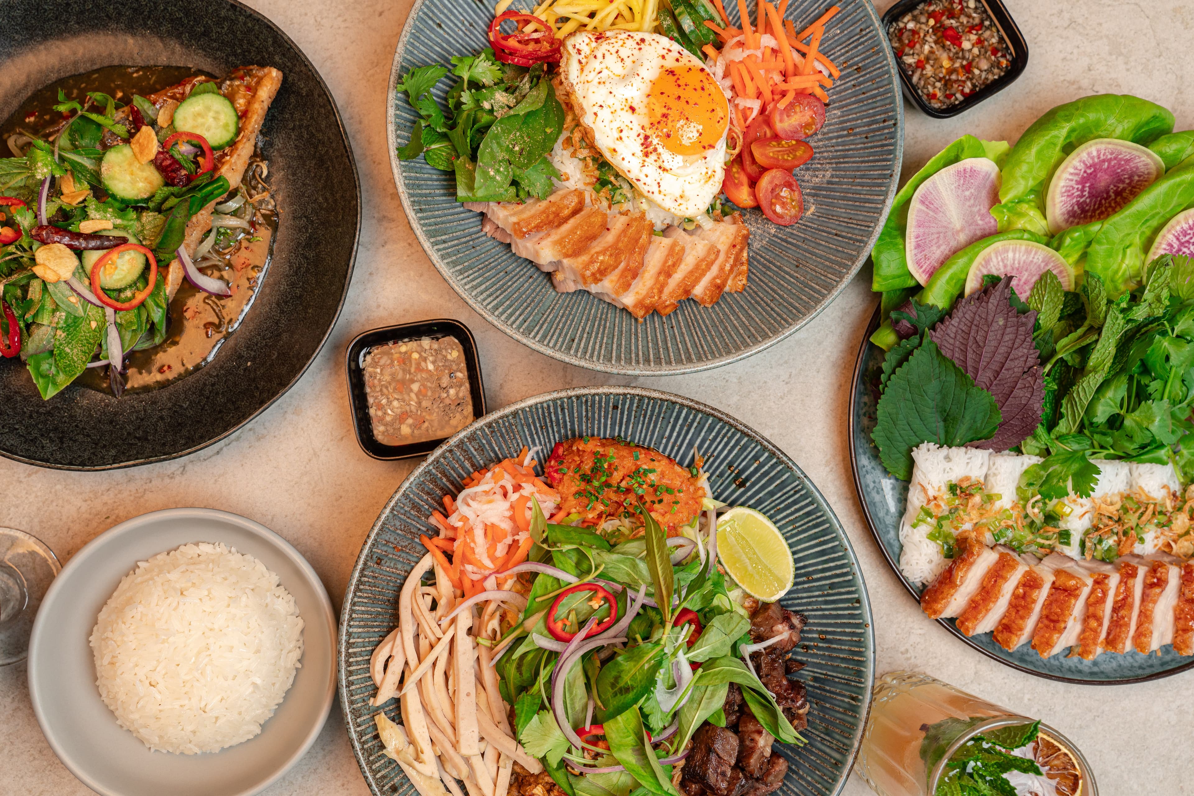 An overhead spread from NEP Cafe featuring a Vietnamese rice bowl with crispy pork belly and sunny-side egg, bun bo hue dry noodles topped with fresh herbs and vegetables, crispy pork belly wraps with rice vermicelli and greens, steamed rice, and sauces.