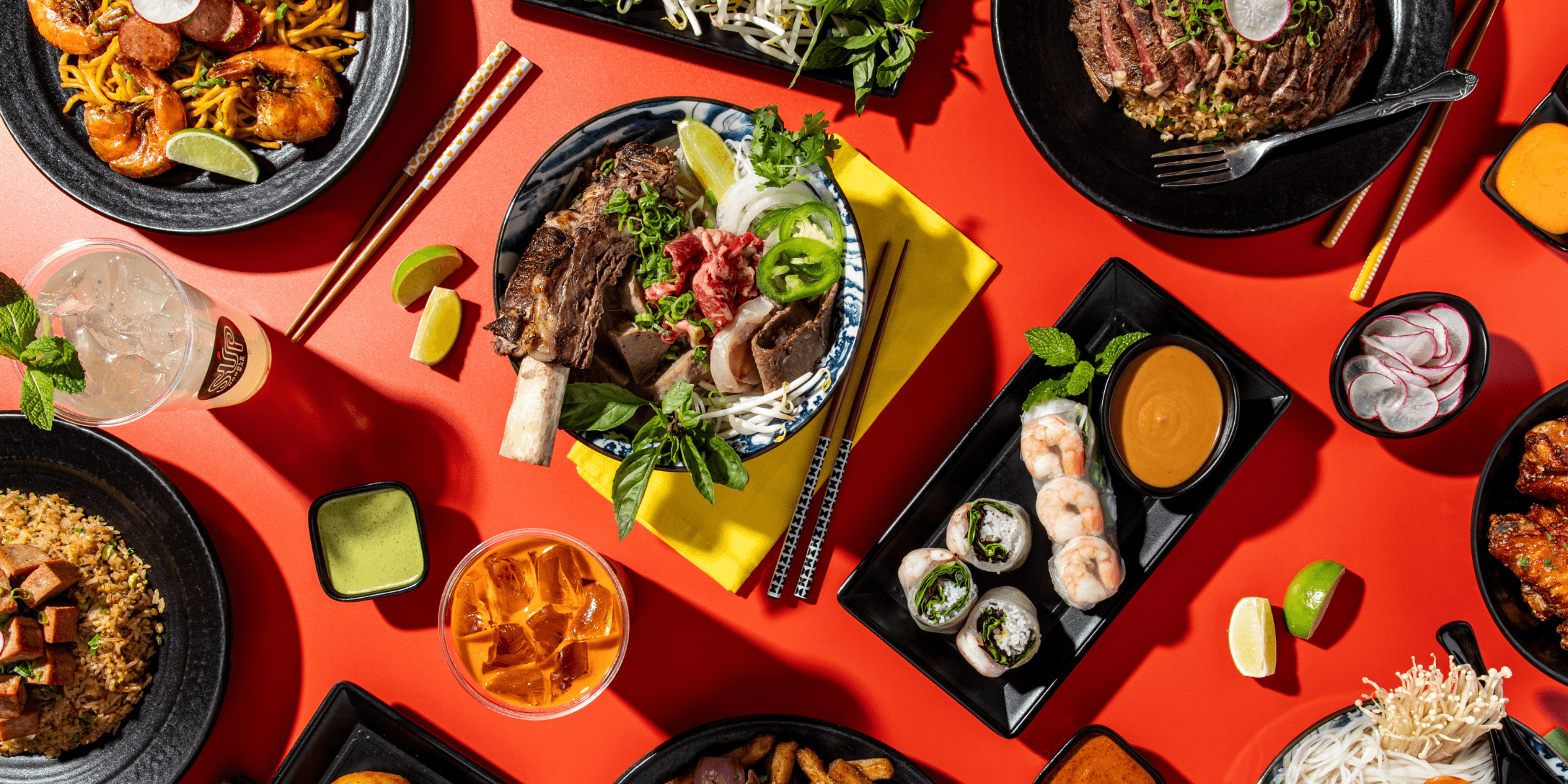 An overhead spread from SUP Noodle Bar featuring combination pho with fresh herbs, shrimp spring rolls with dipping sauce, savory lomo saltado, spam fried rice, and other Vietnamese-inspired favorites arranged across a bright red table.