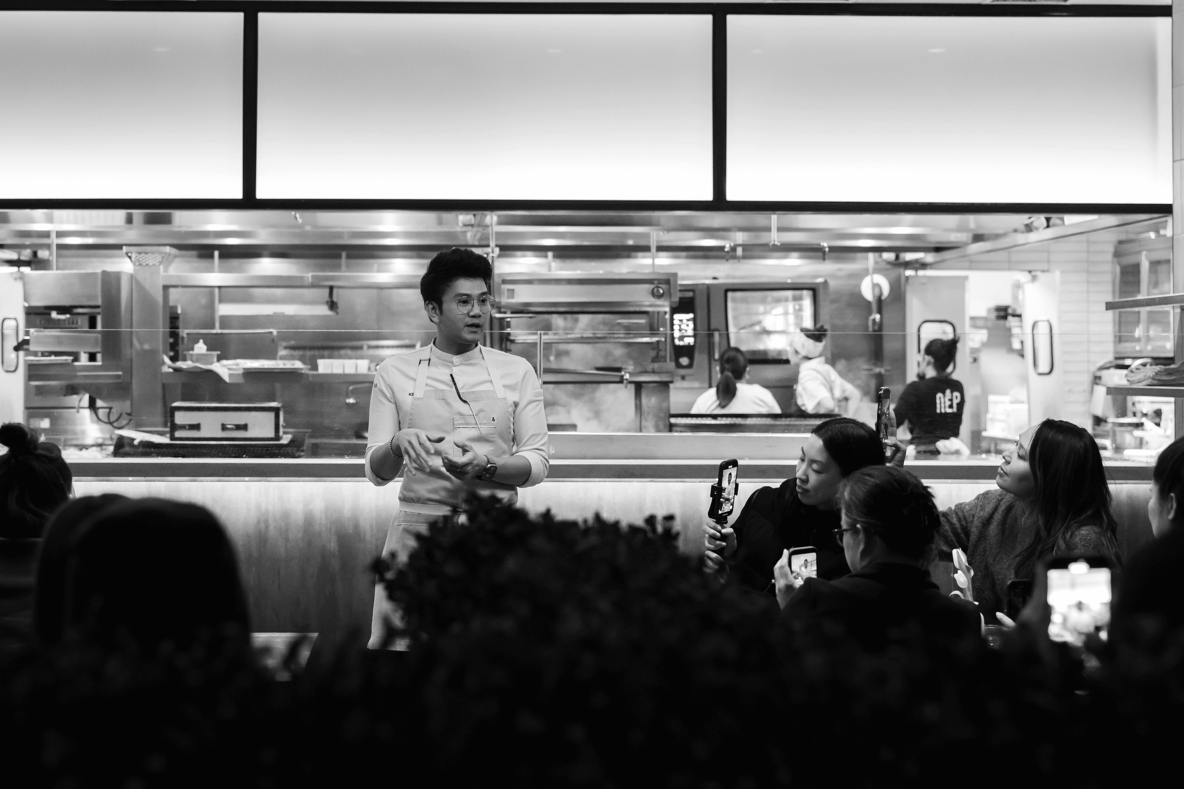 Black-and-white photo from Kei Supper Club featuring a chef presenting dishes to guests during an intimate reservation-only dining experience inside an open-kitchen restaurant setting.