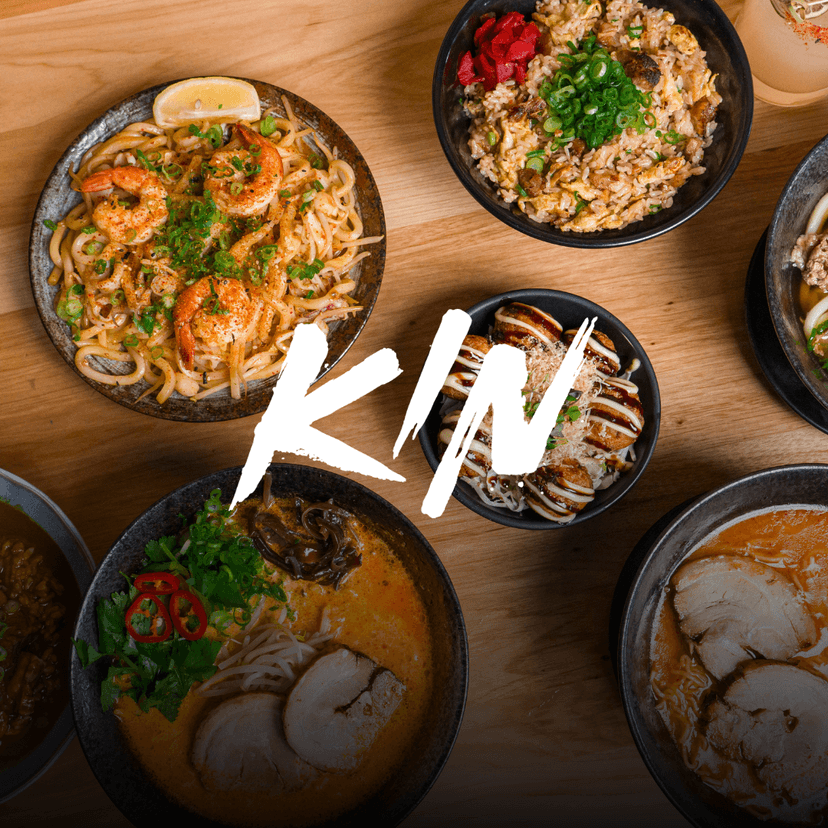 KIN Craft Ramen & Izakaya white logo overlaid an overhead spread from Kin Craft Ramen & Izakaya featuring tom kha ramen, beef belly curry, takoyaki topped with sauce and bonito flakes, beef udon, shrimp garlic noodles, fried rice, and assorted ramen bowls arranged across a wooden table.