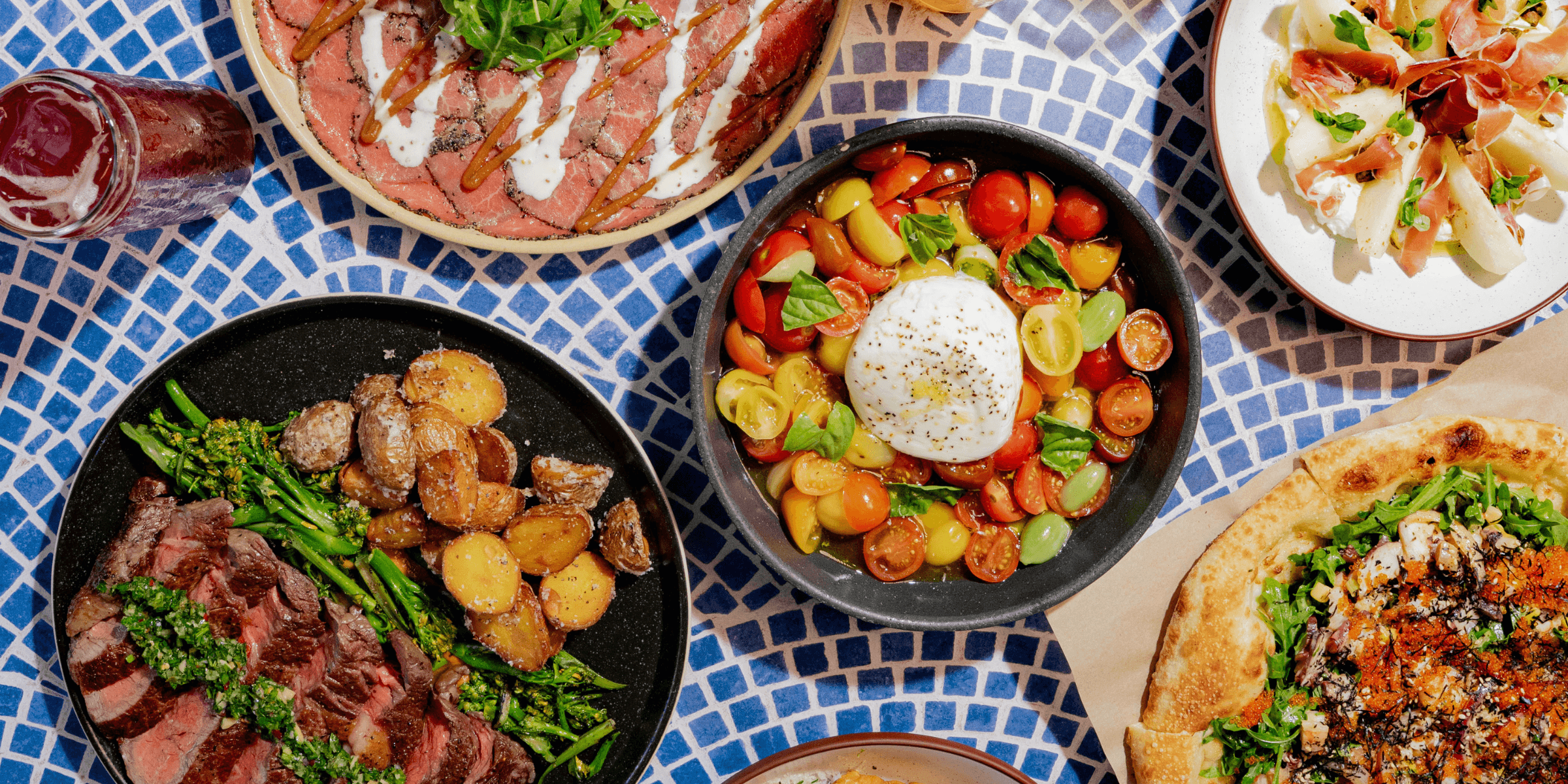 An overhead spread from INI Ristorante featuring burrata caprese with heirloom tomatoes and basil, miso picanha steak with roasted potatoes and broccolini, beef carpaccio topped with arugula and sauce drizzle, pizza, and assorted Italian-inspired dishes arranged across a blue mosaic table.