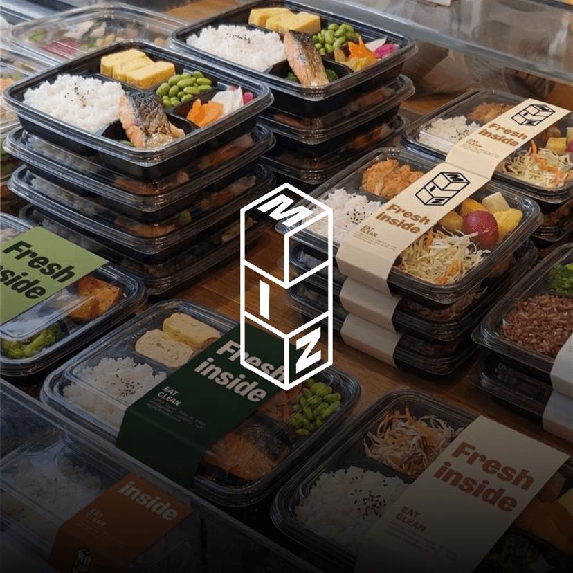 MIZ logo overlaid a stacks of fresh prepared meal boxes arranged on a wooden table, featuring balanced grab-and-go meals with rice, vegetables, fresh fruit, and protein-packed entrées in branded packaging.