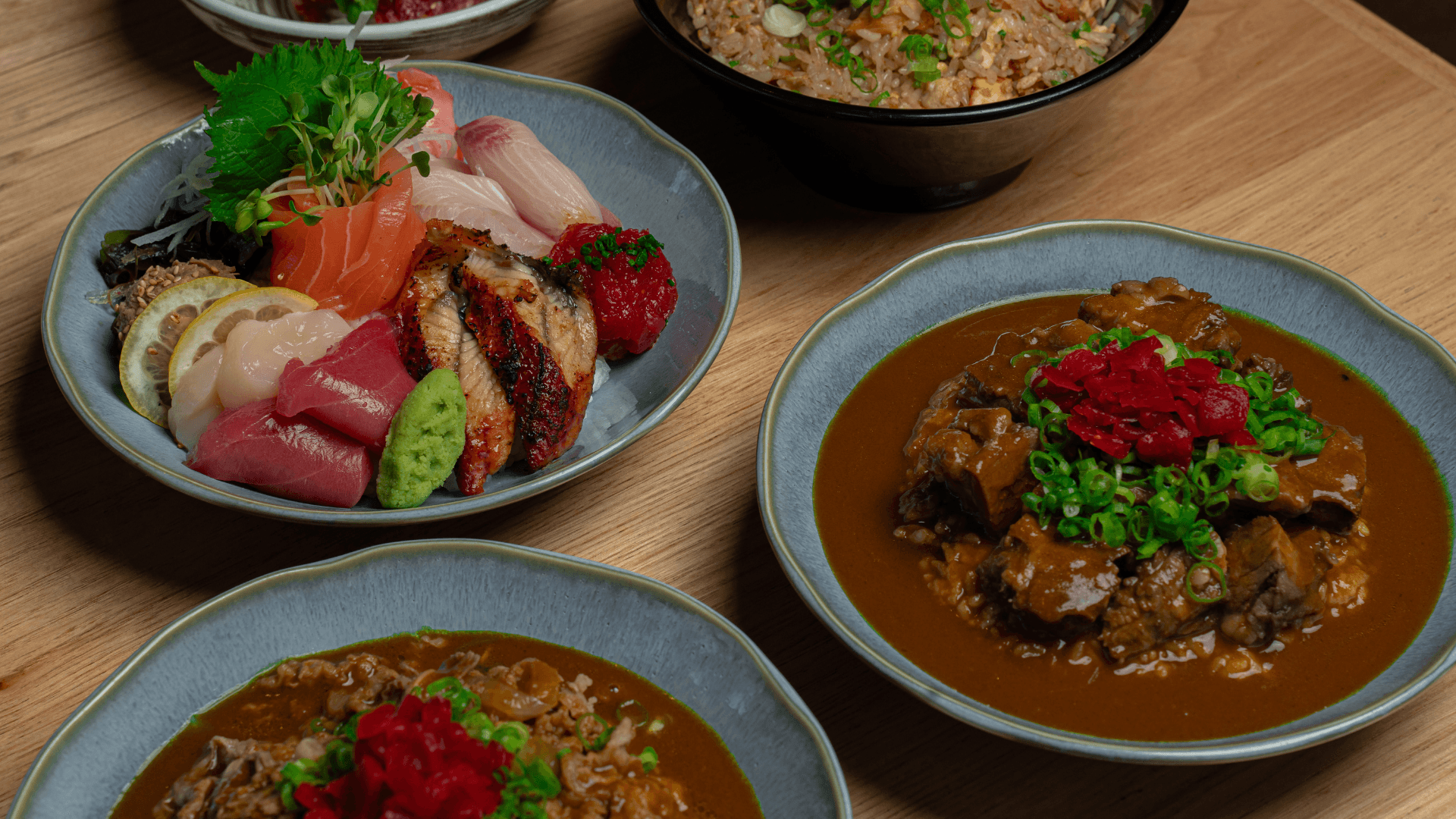A spread from Kin Craft Ramen & Izakaya featuring a chirashi bowl with assorted sashimi, beef curry rice, beef belly curry, and fried rice arranged across a wooden table.