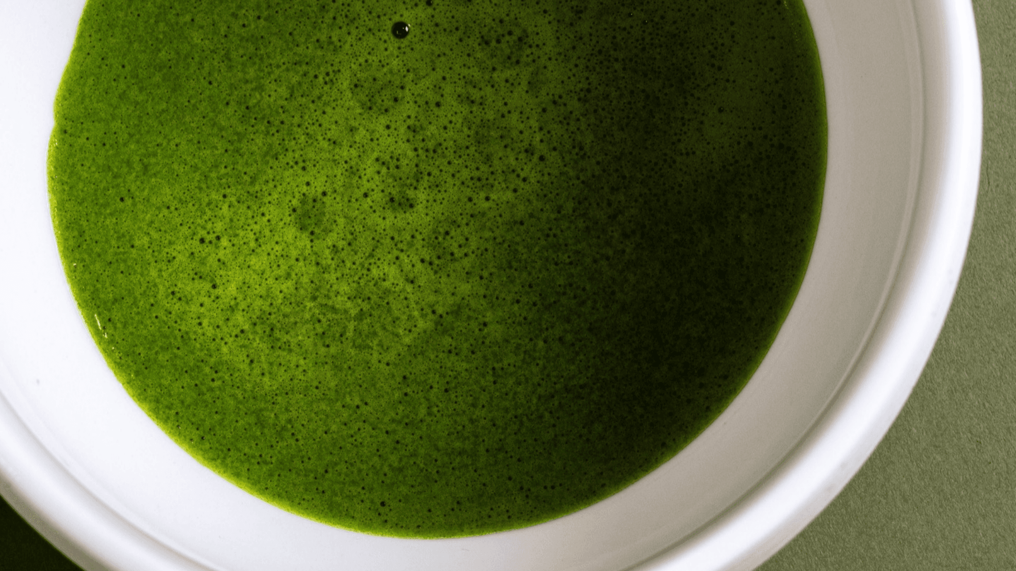 Overhead close-up of vibrant green matcha tea with a fine, frothy surface in a white ceramic bowl against a muted green background.