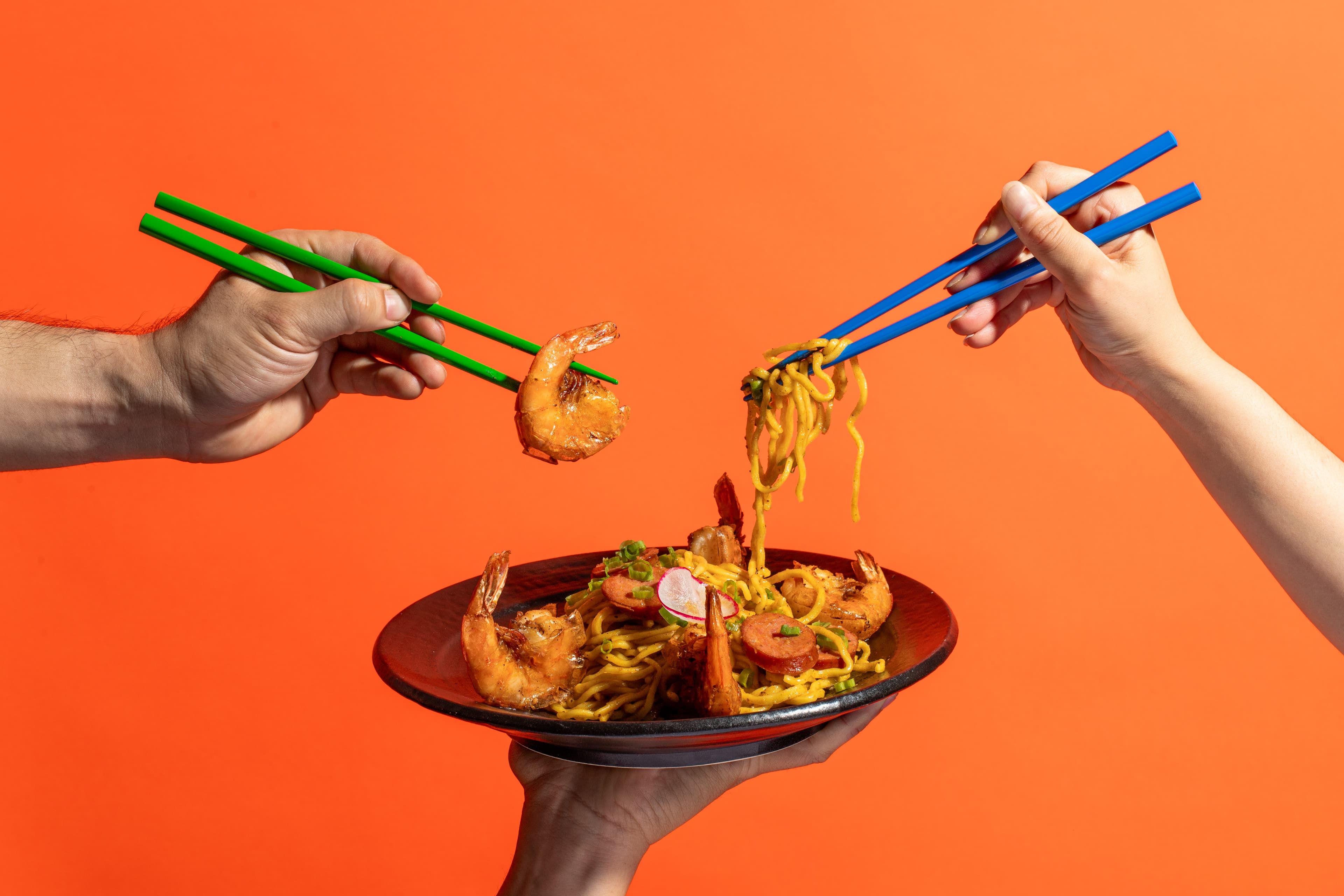 Hands using colorful chopsticks to lift shrimp and sausage Cajun garlic noodles from a bowl at SUP Noodle Bar, featuring grilled shrimp, sausage slices, and garnishes against a bold orange background.